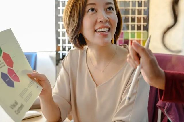 Aasian nainen toimistossa, kirjoittaa paperille kynällä, ympärillä printtimediaa.